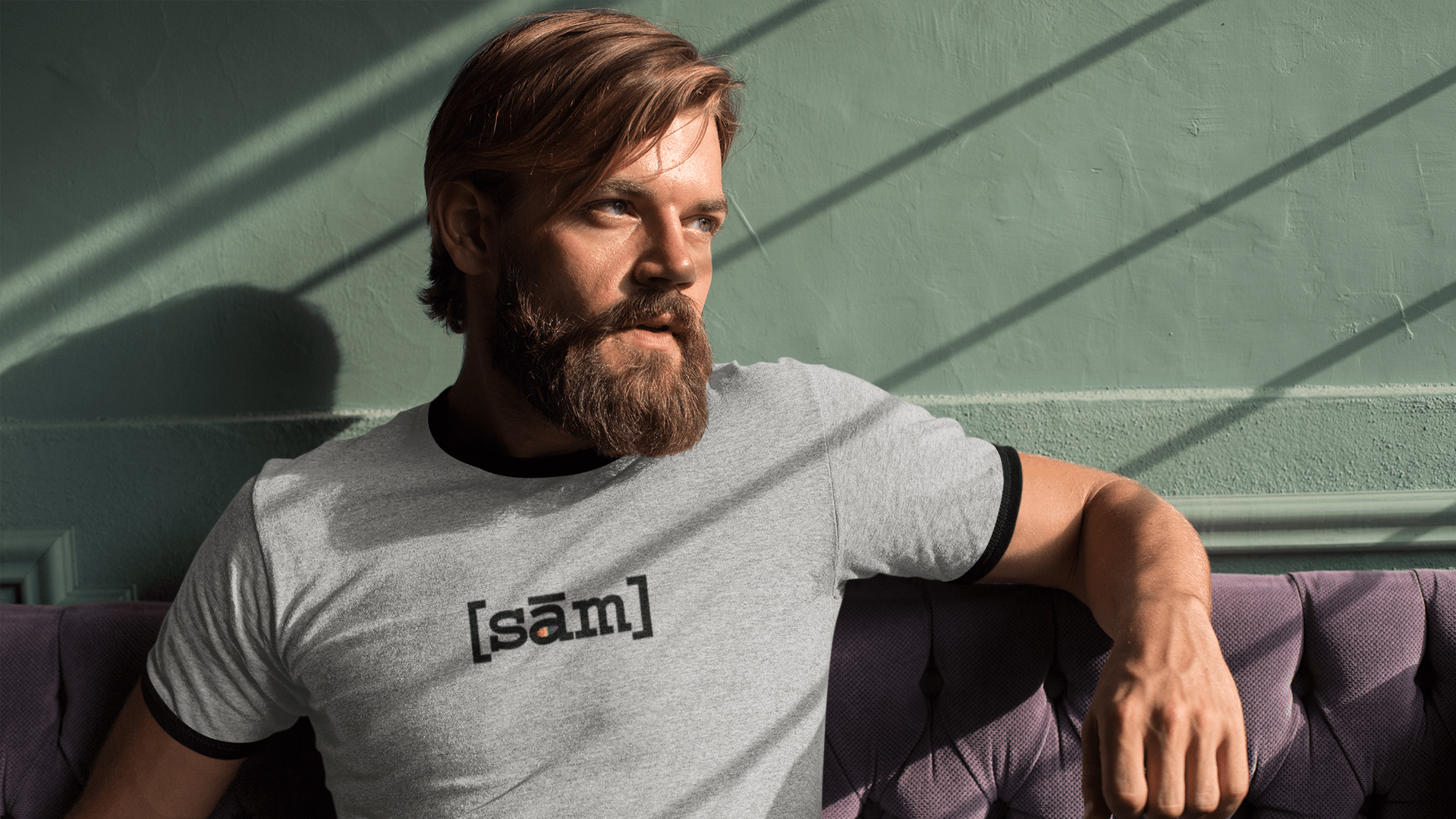 Bearded man sitting on a purple velvet couch with light from a window coming through on his left side. He has his left arm up on the top of the couch. He is wearing an LGBTQ pride t-shirt with the Same Apparel LGBTQ clothing brand logo on the front center of his heather grey vintage ringer shirt.