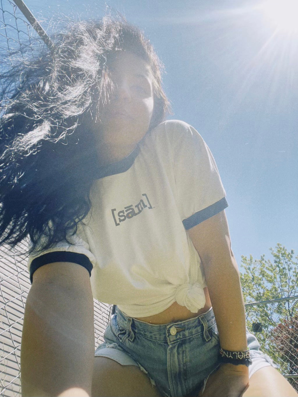 Woman with long black hair to the right of her face looking into camera. Her knees are bent in a squat position with her left hand bracing her body. She has a smirk and is wearing a gay pride shirt with the LBGTQ clothing brand Same Apparel logo on the front center. The LGBTQ vintage white and black ringer shirt is tied in a knot on her left, slightly exposing her midriff.