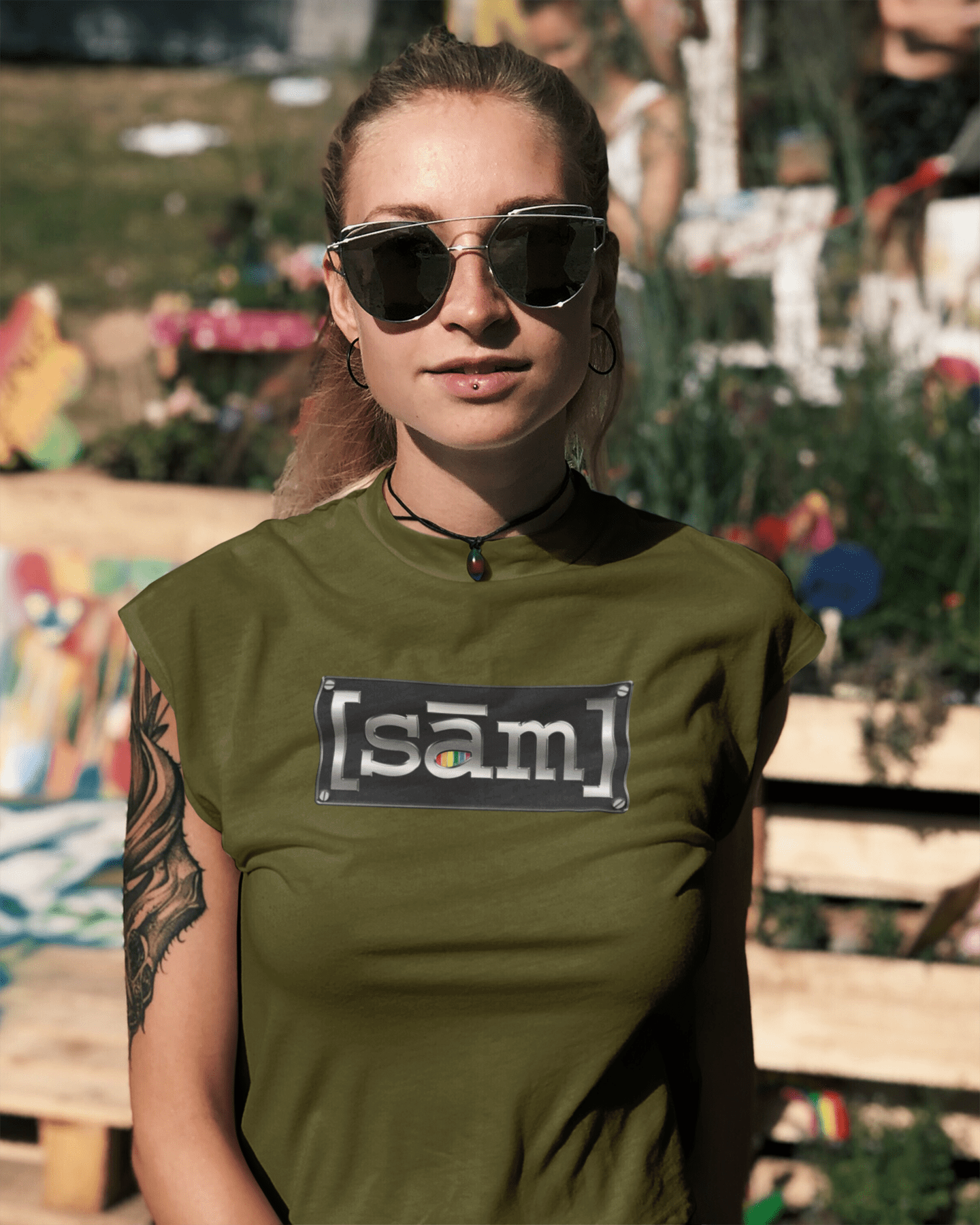 Young woman with lip piercing and arm tattoo, at a party wearing sunglasses and an army green gay pride same apparel muscle t-shirt. The gay same apparel logo is displayed front center of the LGBTQ shirt.