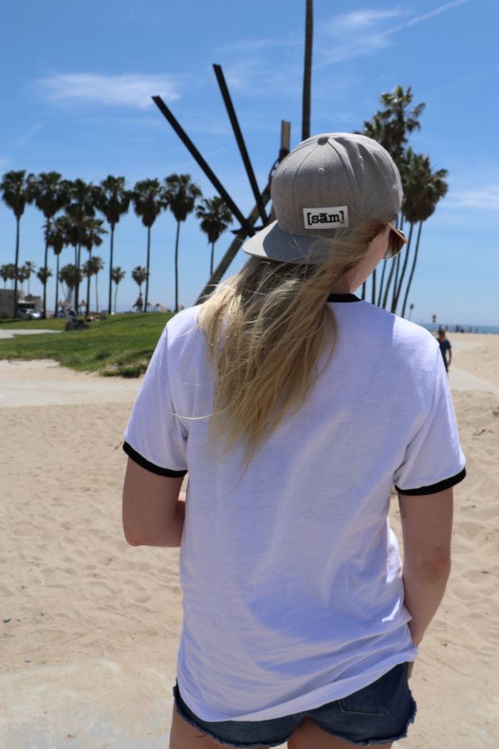Blonde woman with her back facing the camera wearing a gay pride apparel black and white vintage ringer pride t-shirt along with a backwards hat. The LGBTQ hat is a Same Apparel LGBTQ brand flat bill grey hat with the gay apparel LGBTQ clothing brand pride logo in white with black letters.