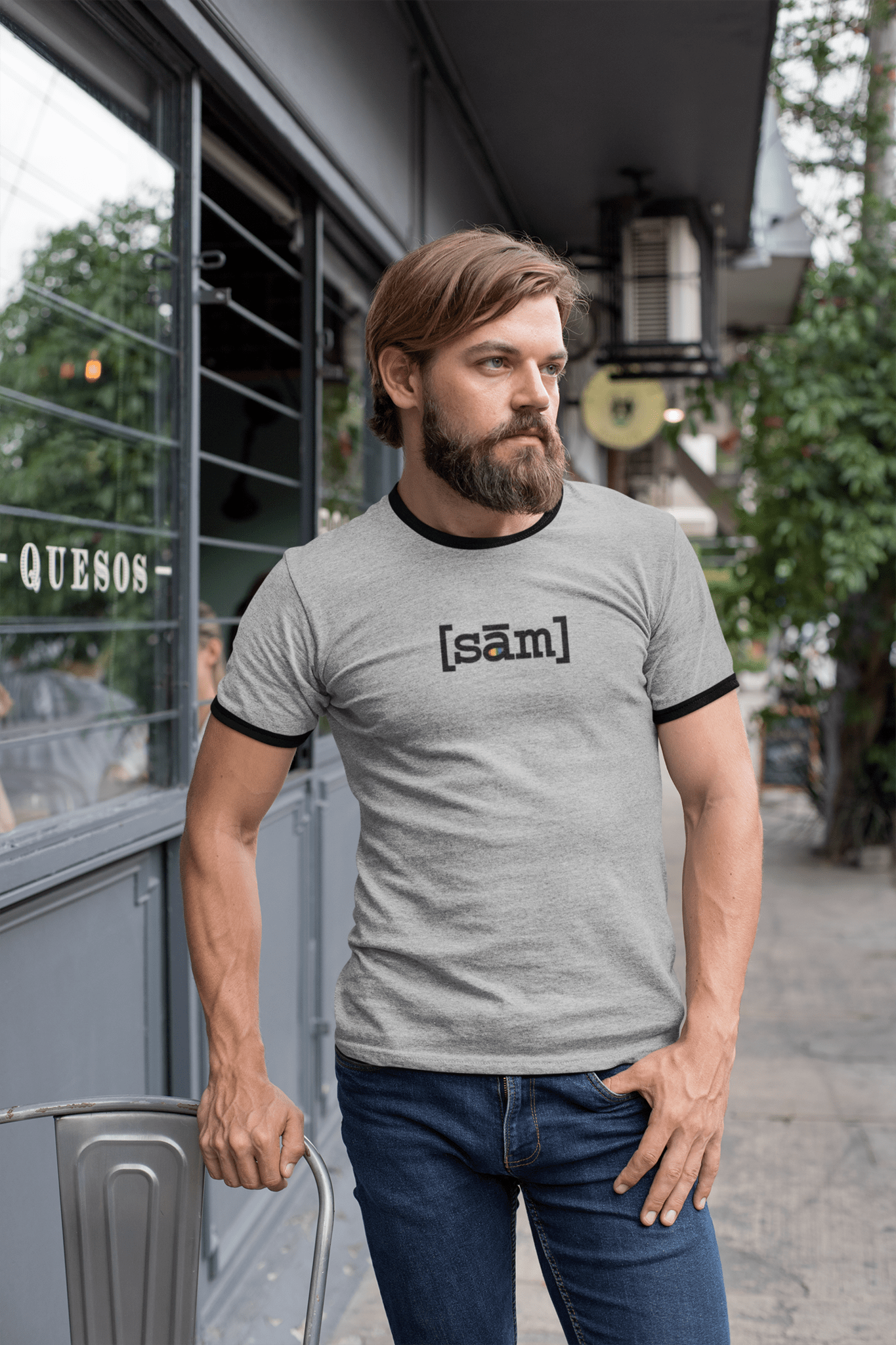 Bearded man standing outside a café with his right hand on the back of a chair and his left thumb in his front jean pocket. He is wearing an LGBTQ gay pride vintage heather grey ringer same apparel clothing brand t-shirt. The gay clothing brand’s logo is on the front center of the pride shirt. 