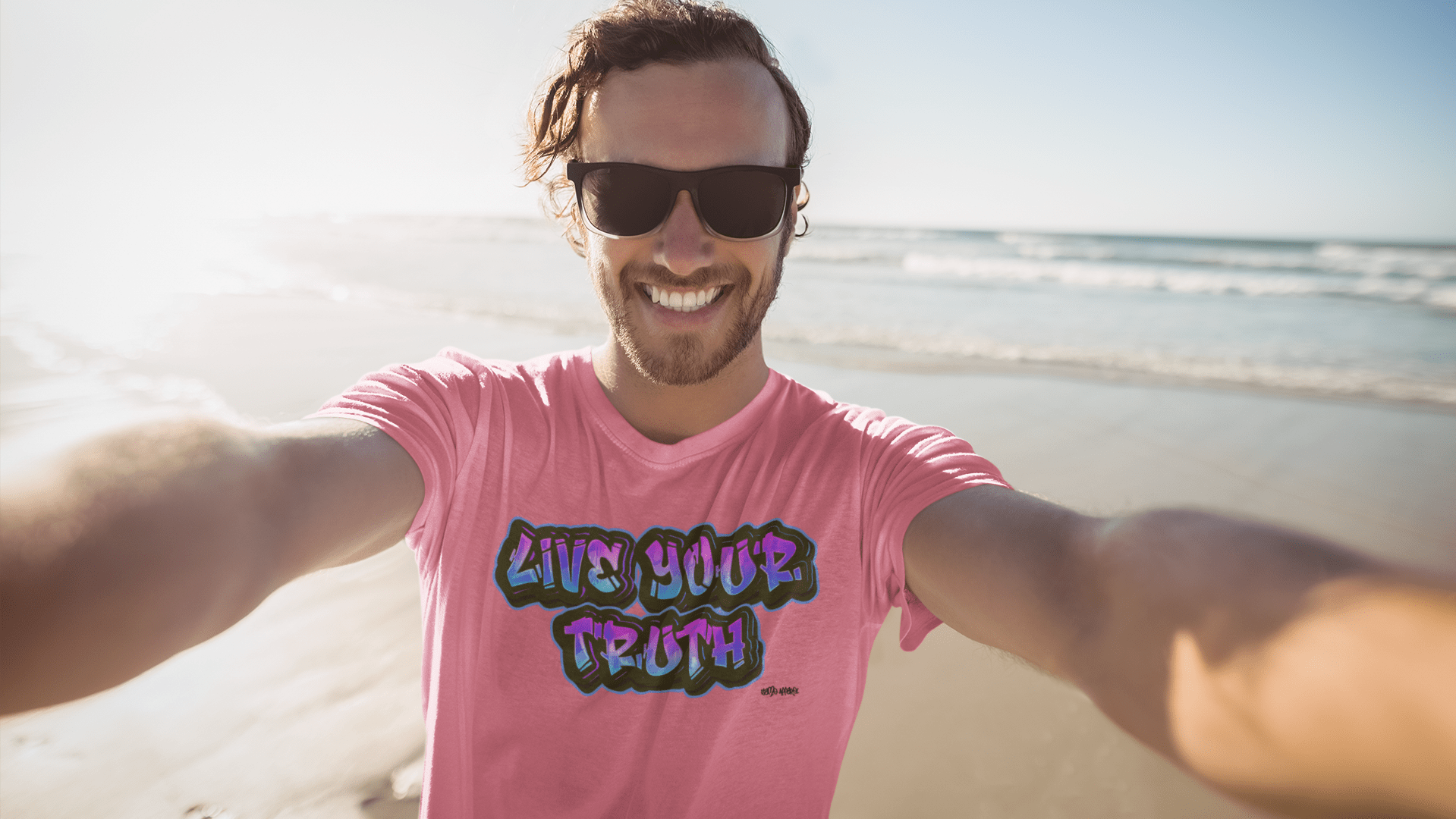 Man standing on a beach with the ocean behind him. He is looking into the camera taking a selfie with both arms extended out taking the picture. He has on sunglasses and is smiling with the sun behind him coming from the left. He is wearing a pink gay pride t-shirt from Same Apparel LGBTQ clothing brands Live your truth gay pride t-shirt collection. The graphic is graffiti style text with "Live your truth" written on the front center of the pride shirt.
