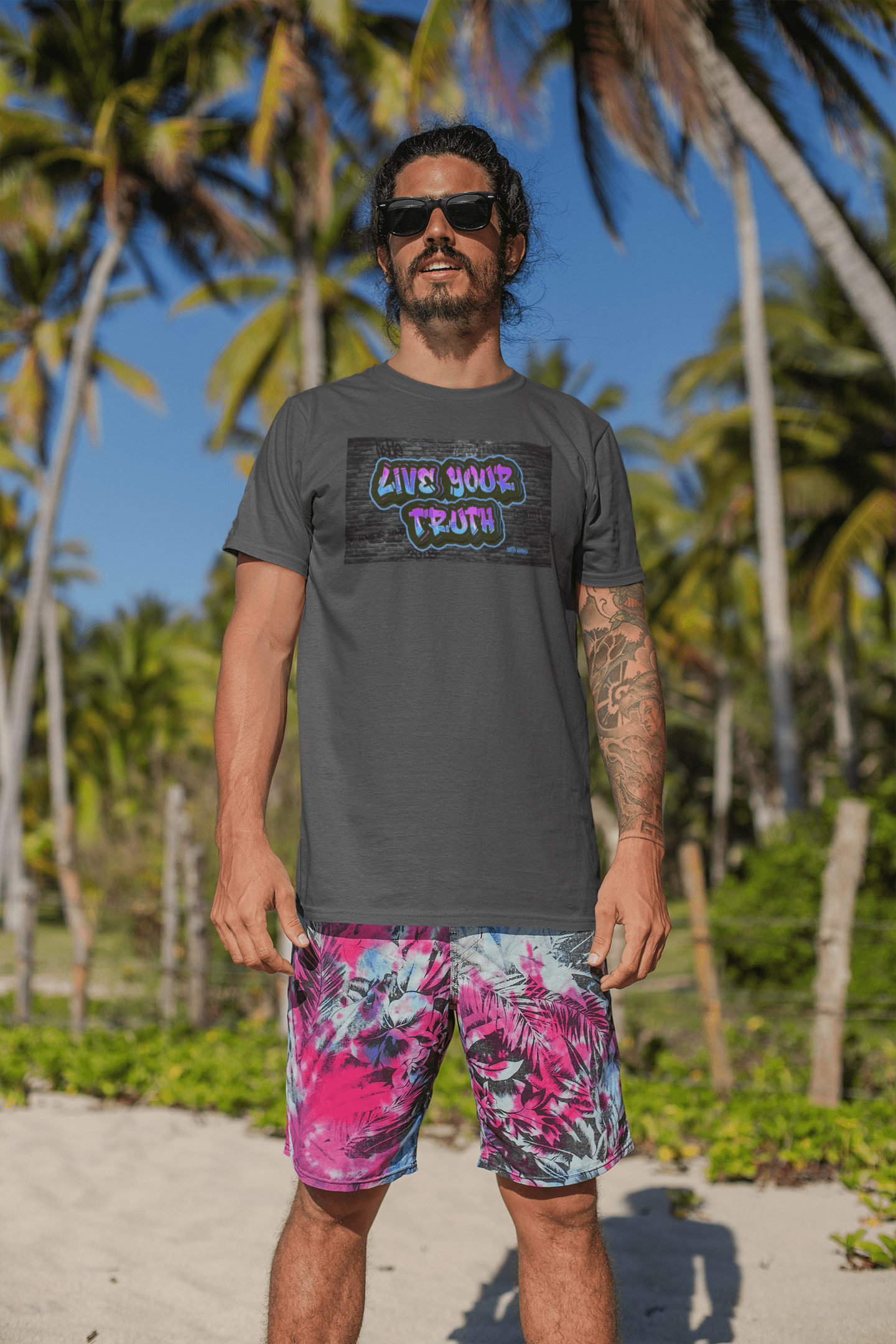 Dark haired man standing on a beach with palm trees in the back of him. He is facing the water, it’s a sunny day. He is wearing black sunglasses with a gay pride t-shirt that has a front graffiti graphic that says “Live your truth.” The LGBTQ shirt is from Same Apparel gay clothing brand.